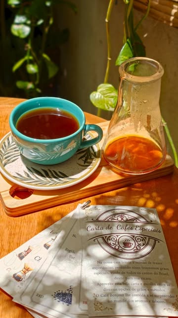 Café coado em xícara azul-turquesa ao lado de uma carafa de vidro, com a Carta de Cafés Especiais sobre a mesa de madeira ao sol.