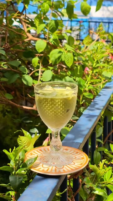 Taça de vidro com bebida gelada de tom claro sobre um pratinho laranja, na grade azul da varanda.