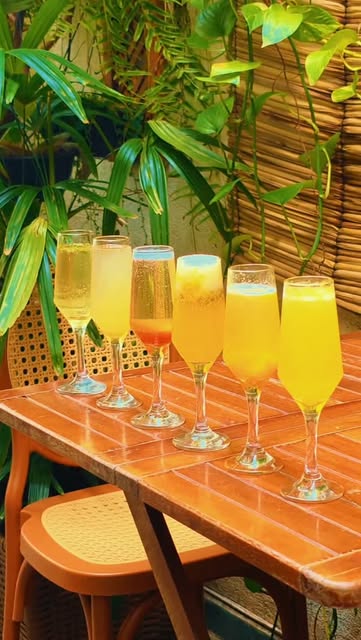 Seis taças de drinks com espumante em fila sobre uma mesa laranja, com plantas ao fundo.