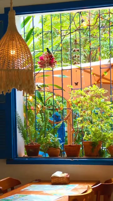 Janela com esquadria azul-cobalto, luminária de palha e vasos de plantas no peitoril, sobre uma mesa de madeira.